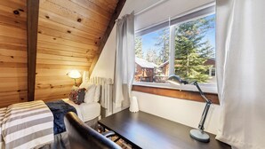 Ein gemütliches Zimmer mit Holzbalkendecke, einem Bett mit gestreifter Bettwäsche, einem Schreibtisch mit Lampe und einem Fenster mit Vorhängen.