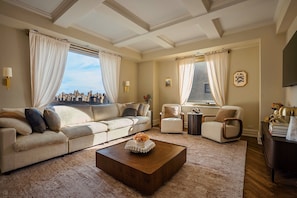 A spacious living room with a large window offering a city view, a beige sofa, a wooden coffee table with a decorative vase, and a classic ceiling design. - New 2BR in Heart of Manhattan Central Park Views (New York)