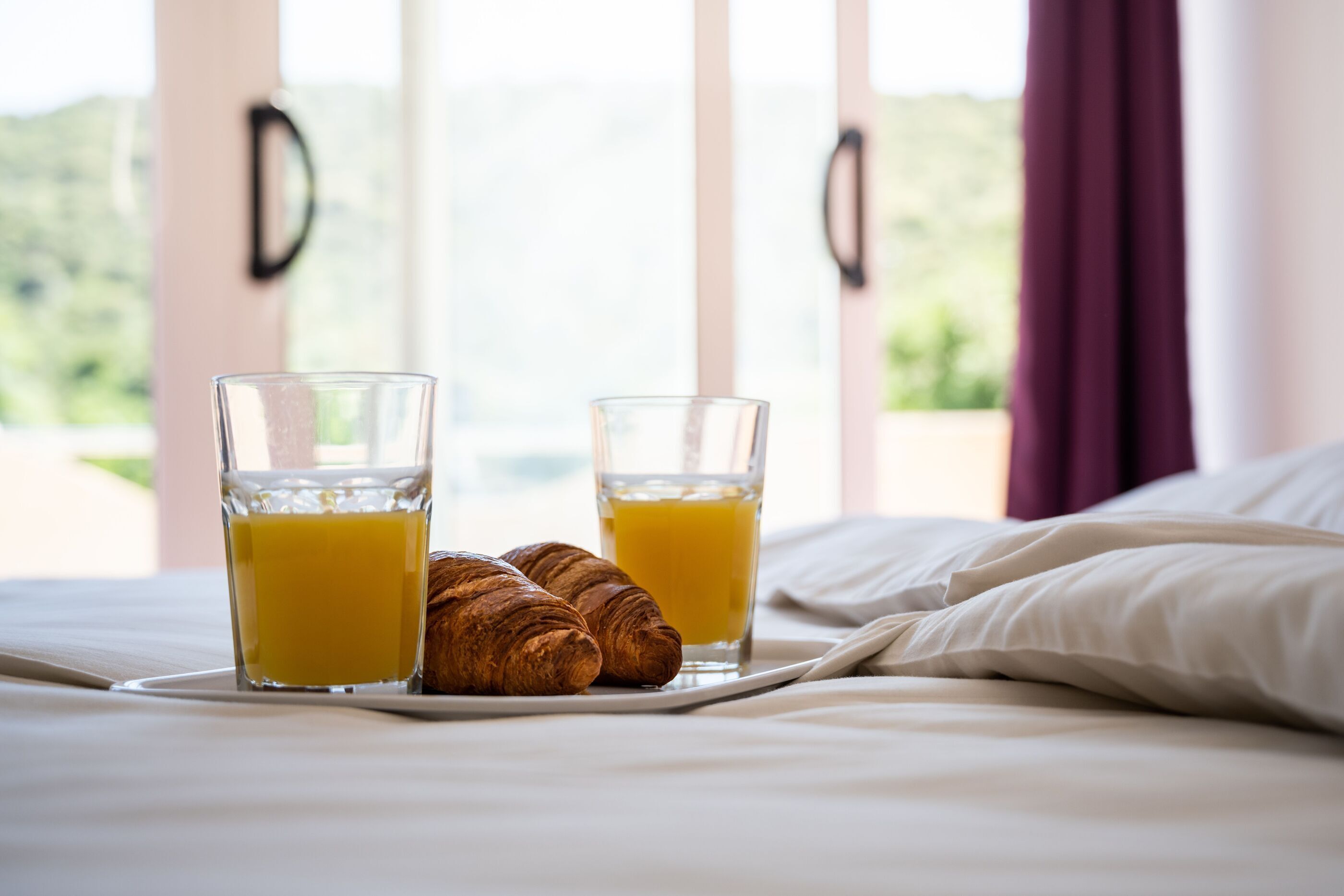 A bed with a tray of croissants and two glasses of orange juice, with a view of greenery through the window.