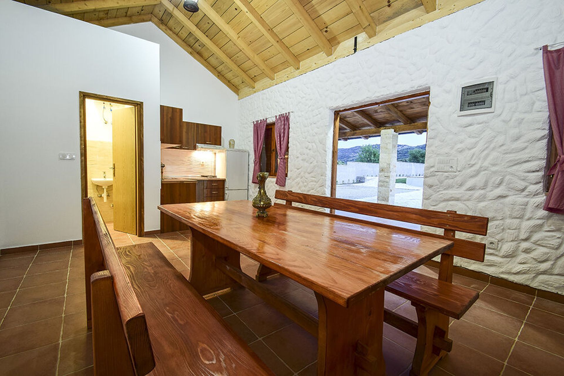 Una mesa de madera con bancos, una zona de cocina y vistas a través de una ventana.