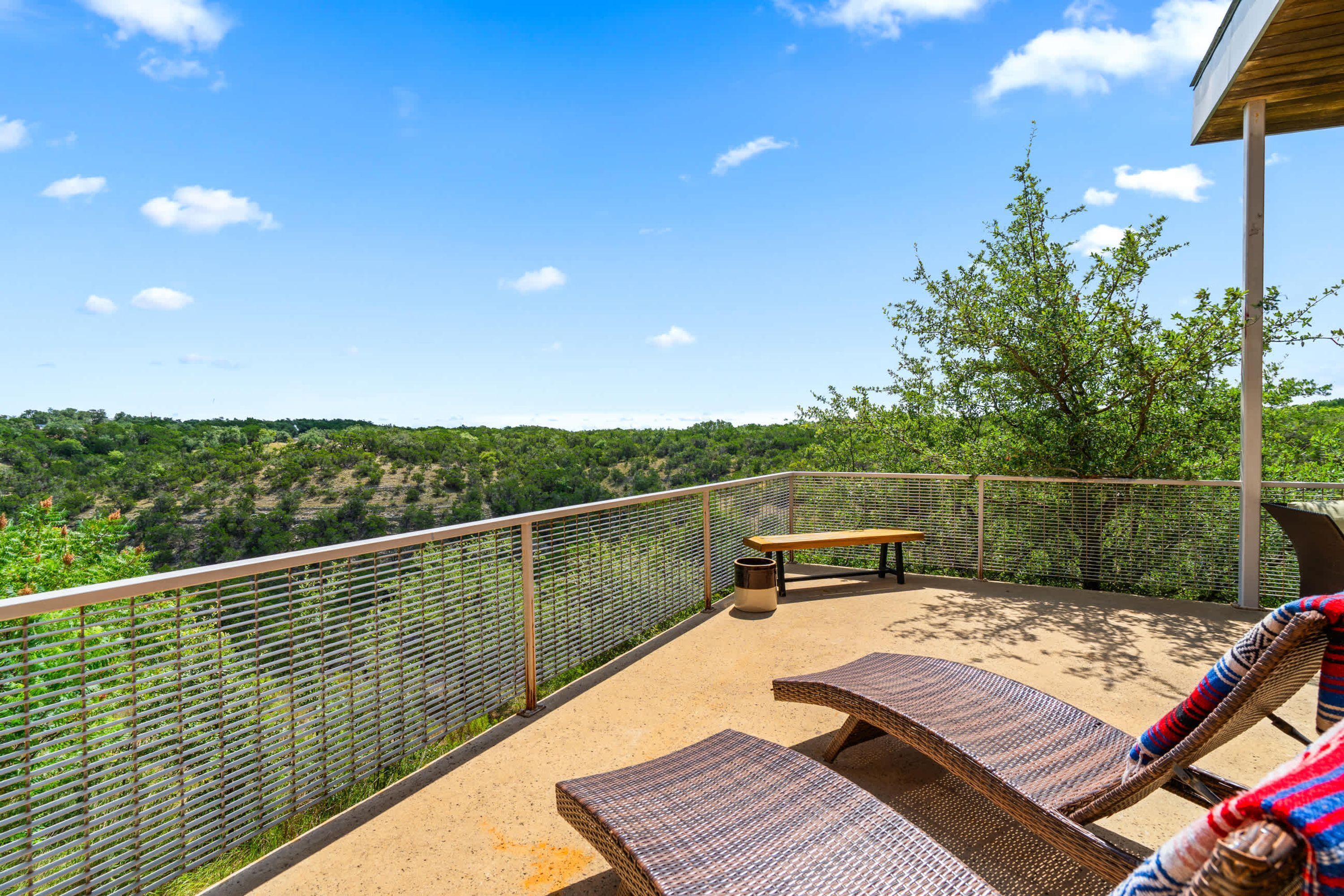 A balcony with wicker lounge chairs, a wooden table, and a metal fence overlooking a forested landscape.