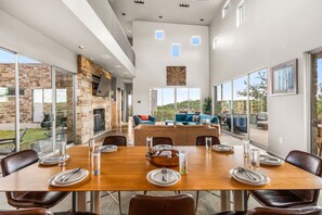 A spacious dining area with a wooden table set for six, surrounded by leather chairs. A stone fireplace and a large window provide a view of the outdoors. - Luxury 4BR Hilltop Retreat With Scenic Views (Dripping Springs)