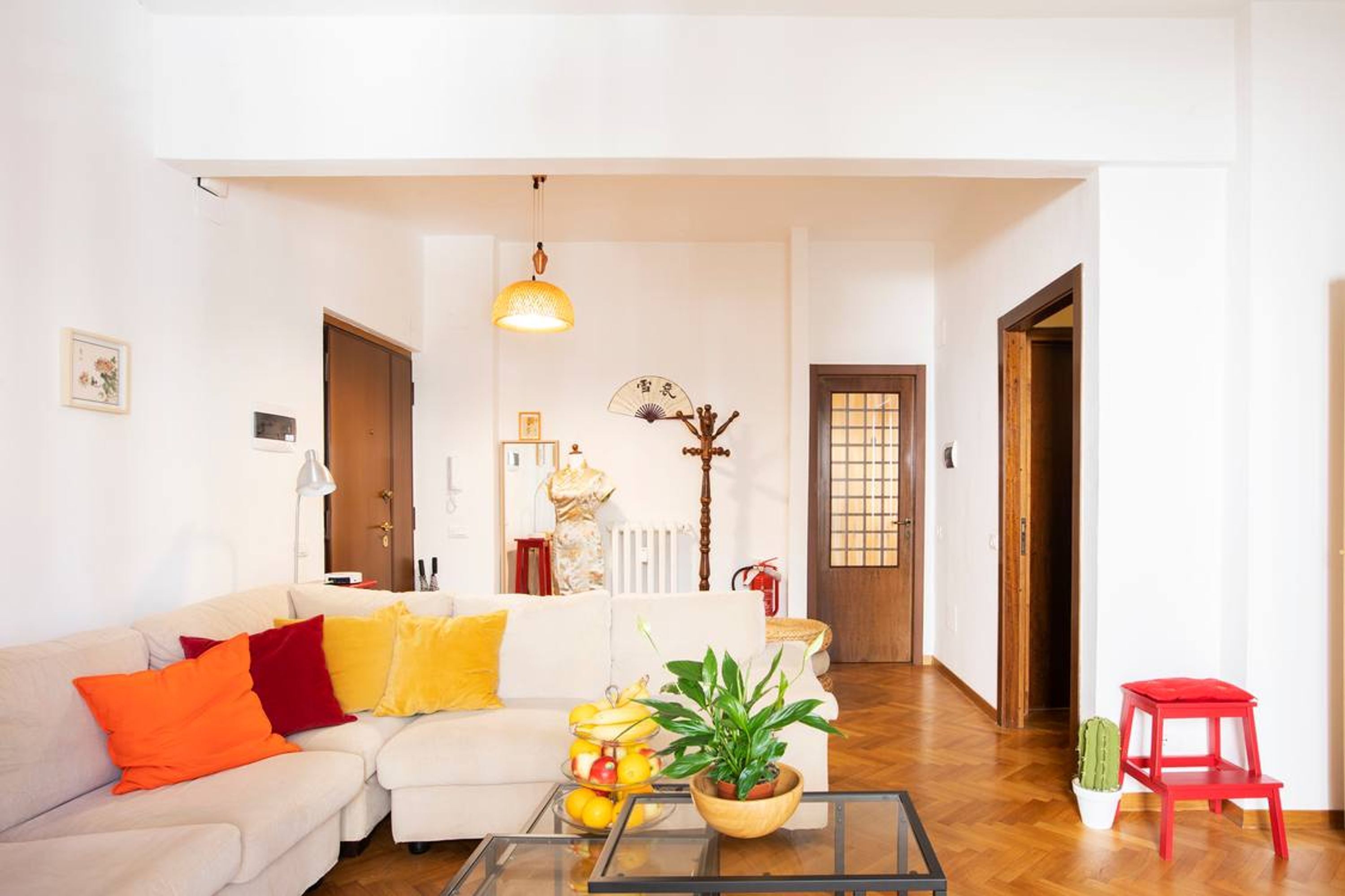 A living room with a sofa, a coffee table with fruits and a plant, a red stool, and a wooden door in the background.