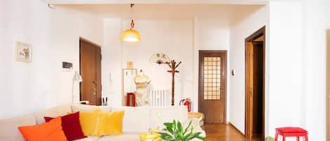 A living room with a sofa, a coffee table with fruits and a plant, a red stool, and a wooden door in the background.