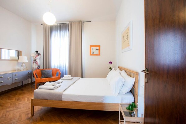 A hotel room with a wooden bed, a bedside table with a lamp, a chair, a window with curtains, and a door. - Casa Romitino Modern and Spacious Apartment (Florence)