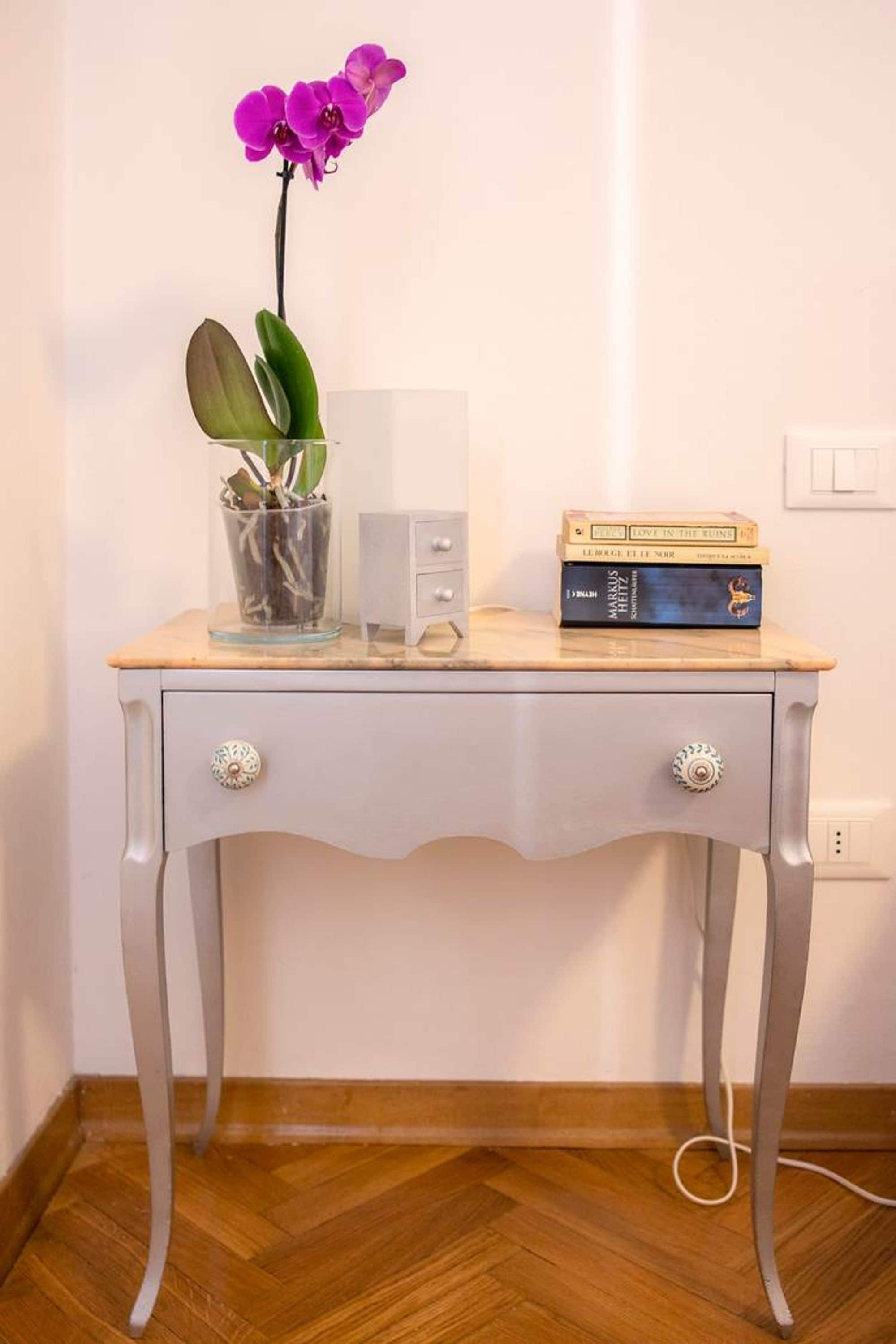 Un petit bureau blanc sur lequel se trouvent un vase en verre contenant une orchidée violette et une plante en pot, une pile de livres et un tiroir blanc.