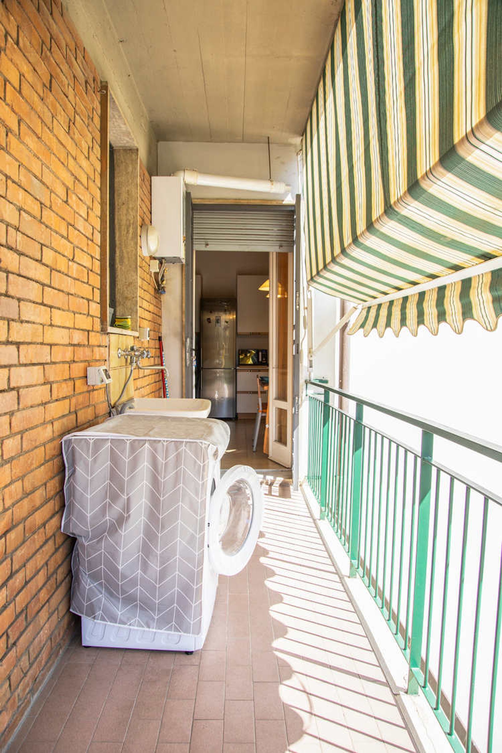 A balcony with a covered outdoor toilet, a brick wall, a kitchen area, and a striped awning.