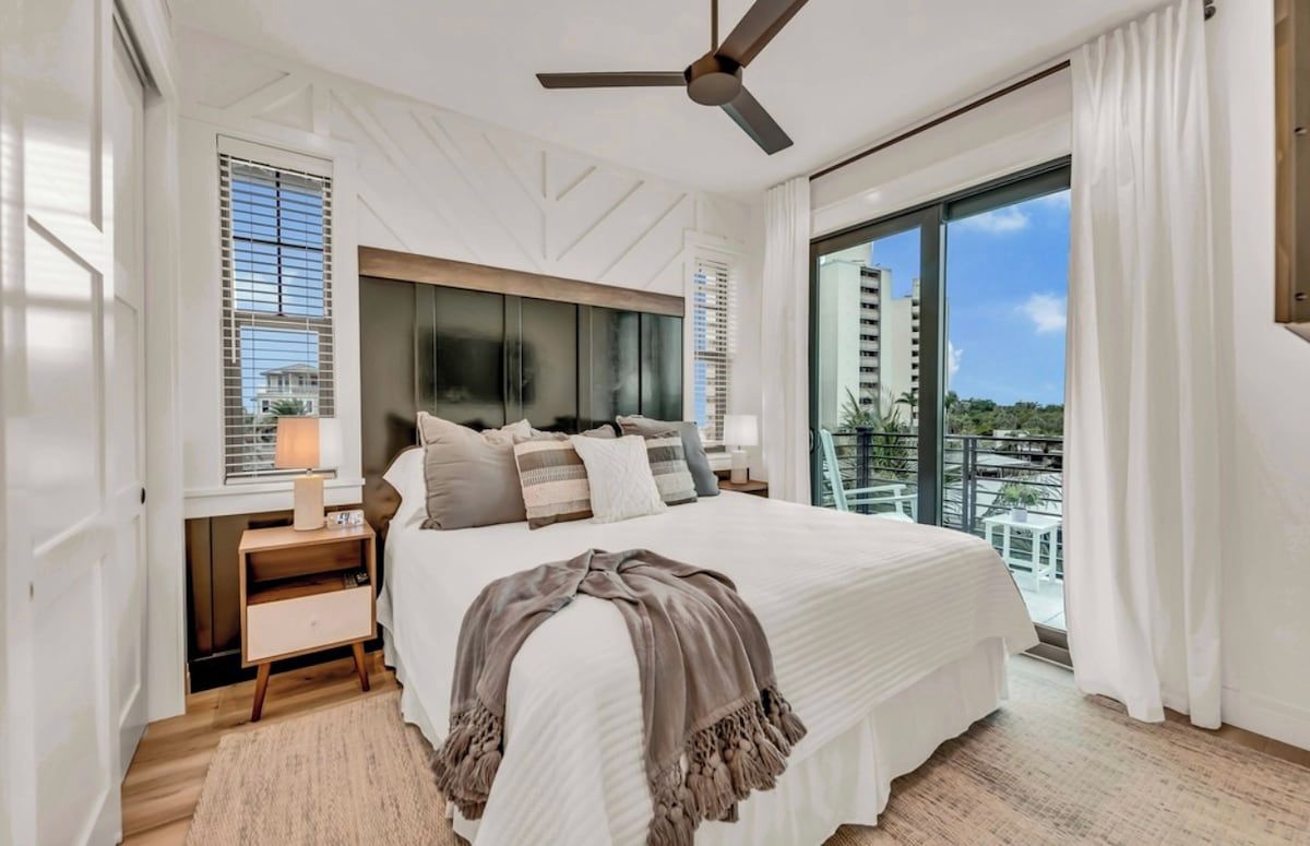 Une chambre moderne avec un grand lit, un ventilateur de plafond et une vue sur le paysage urbain, visible à travers une grande fenêtre.