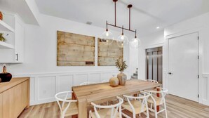 Una cocina moderna con una mesa de comedor de madera, sillas blancas, iluminación colgante y un cuadro abstracto grande en la pared.
