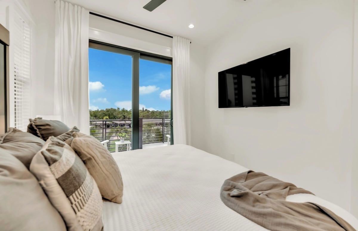 A bedroom with a large bed, a flat-screen TV mounted on the wall, and a balcony with a view of greenery.