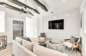 Ein modernes Wohnzimmer mit einer Couch, Sesseln, einem an der Wand montierten Fernseher und einem Deckenventilator.