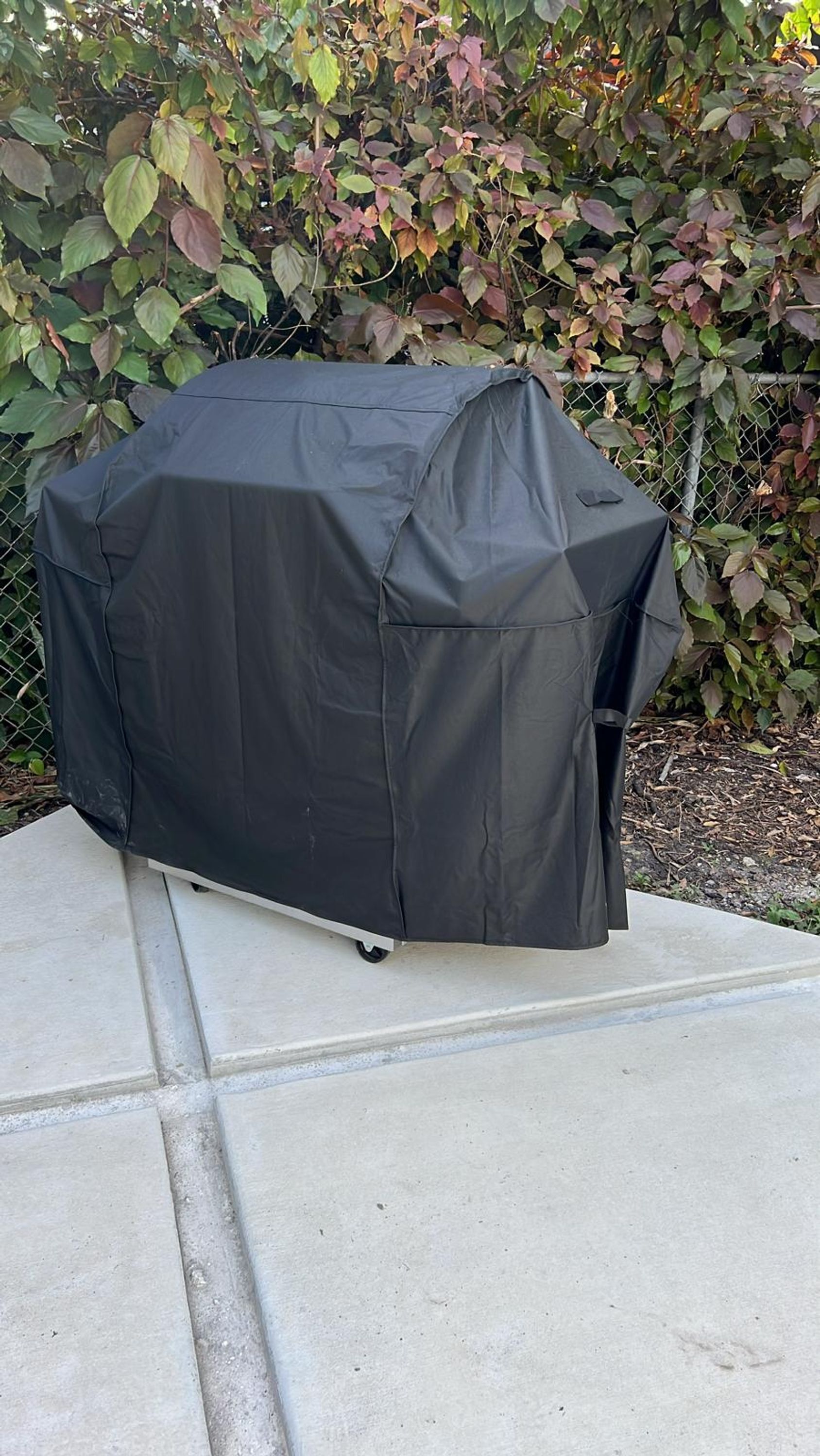 A covered grill on a concrete patio with a chain-link fence and foliage in the background.