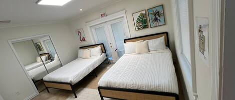A bedroom with two single beds, a large mirror, a door with an exit sign, and two framed tropical leaf paintings.