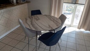 Un comedor con una mesa de mármol y dos sillas, una encimera de cocina con cafetera, y vistas de un edificio a través de una ventana.