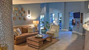 Ein geräumiges Wohnzimmer mit Sofa, Sessel und Couchtisch. An den Wänden sind dekorative Elemente angebracht und ein großes Fenster lässt viel Tageslicht herein.