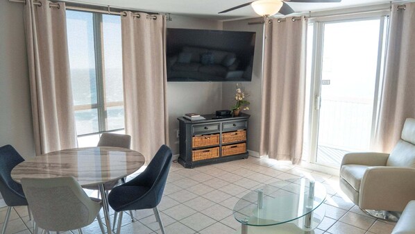 Una sala de estar moderna con una mesa de comedor de vidrio, dos sillas, un sofá, un televisor de pantalla plana montado en la pared y una puerta corrediza de vidrio que da a un espacio exterior.