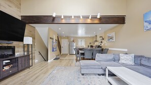 A modern living room with a grey sofa, a coffee table, and a TV on a wooden stand. There is a kitchen area in the background with a dining table and chairs. - Pocono Paradise Ultimate Camelback Getaway 4 Bedroom Townhouse (Tannersville)