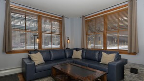 Ein Wohnzimmer mit einem blauen Ecksofa, einem Holztisch und großen Fenstern mit Jalousien.