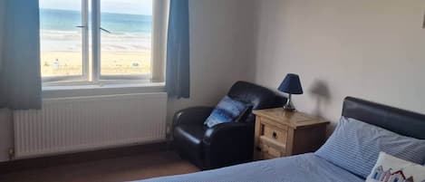 A bedroom with a bed, a chair, a nightstand, and a window overlooking a beach.