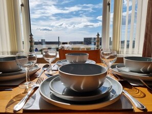 Une table à manger dressée avec des bols, des assiettes et des couverts, offrant une vue sur le paysage urbain à travers de grandes fenêtres.
