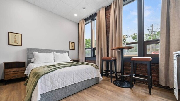 Una habitació d'hotel moderna amb un llit gran, dues nits, una taula rodona amb tamborets i finestres grans amb cortines.