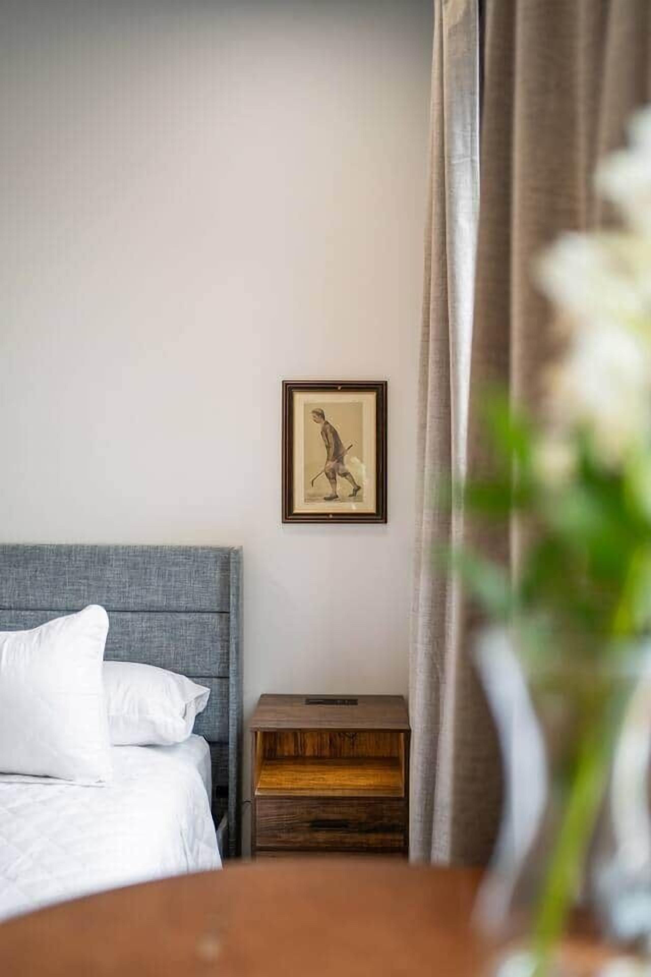 Un dormitorio con cama, mesita de noche, un cuadro enmarcado en la pared y una cortina que cubre parcialmente una ventana.