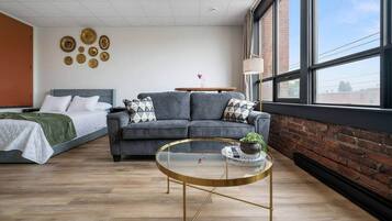 Una habitación de hotel moderna con un sofá gris, una cama con una manta verde, una mesa de centro redonda de vidrio con una planta y una pared de ladrillo.
