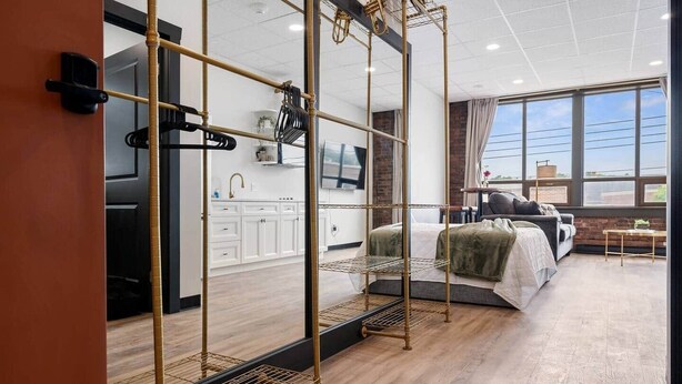 A modern hotel room with a large bed, a TV, a kitchen area, and a view of the city through large windows.