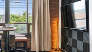 A modern room with a brick wall, a large window with curtains, a round table with stools, and a mounted TV.