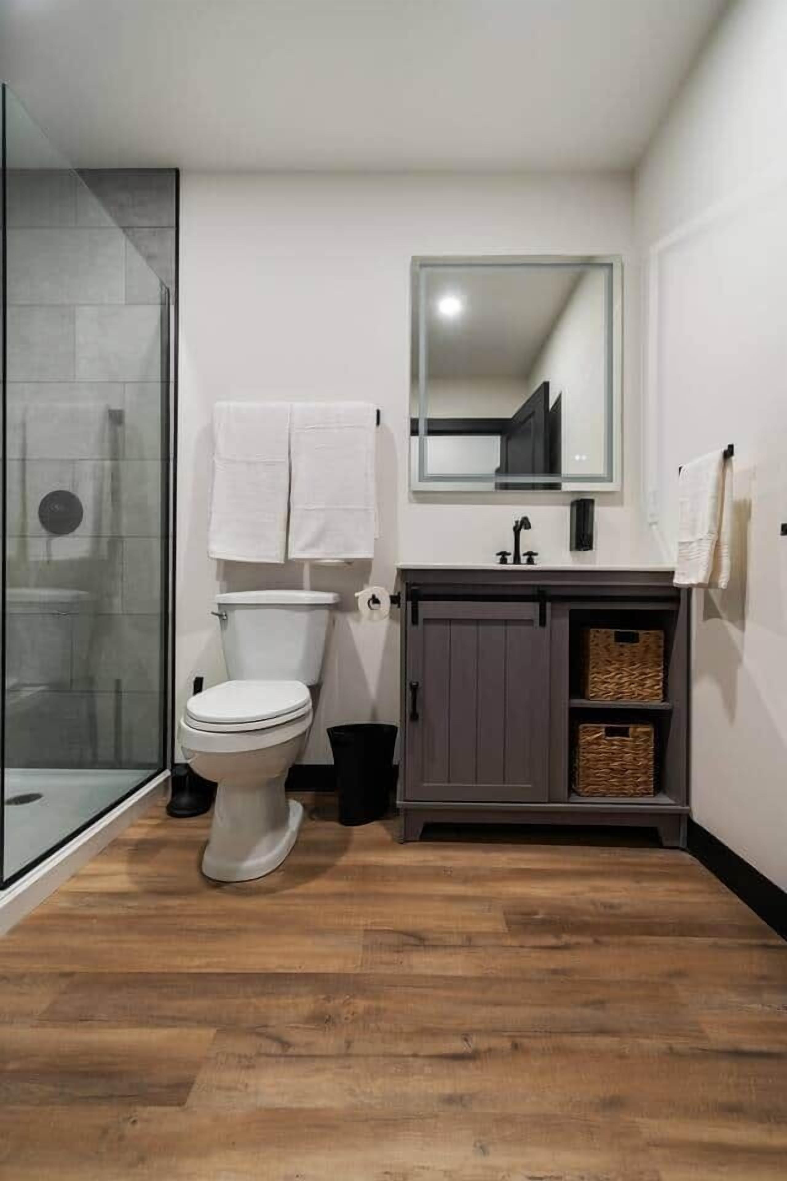 Een badkamer met een douchecabine van glas, een wit toilet, een wastafel met spiegel en een kastje met manden.