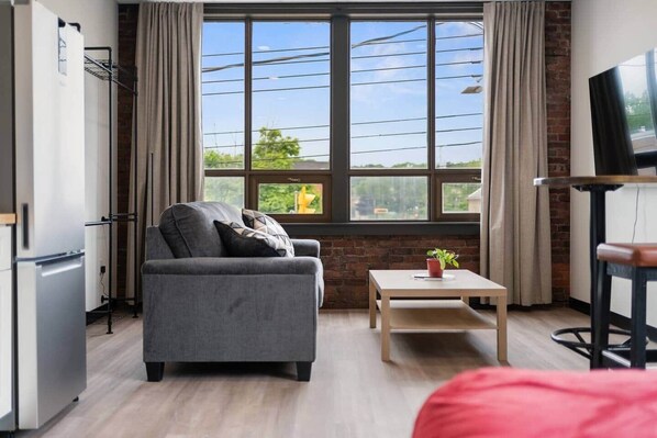A modern living room with a grey sofa, a small table with a plant, a TV on a stand, and a large window with curtains.
