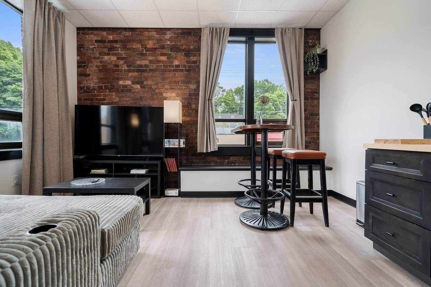 A modern room with a brick wall, a TV, a sofa, a dining table with stools, and a wooden cabinet.