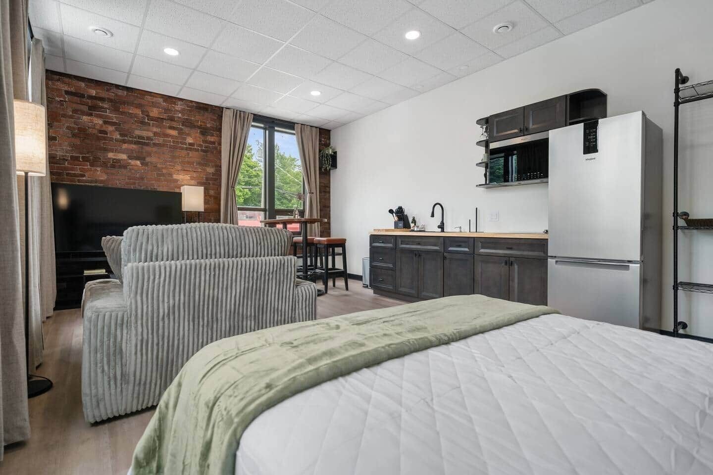 Una habitación de hotel moderna con una cama, un rincón de estar con sillas, una kitchenette pequeña y una pared decorada con ladrillos.