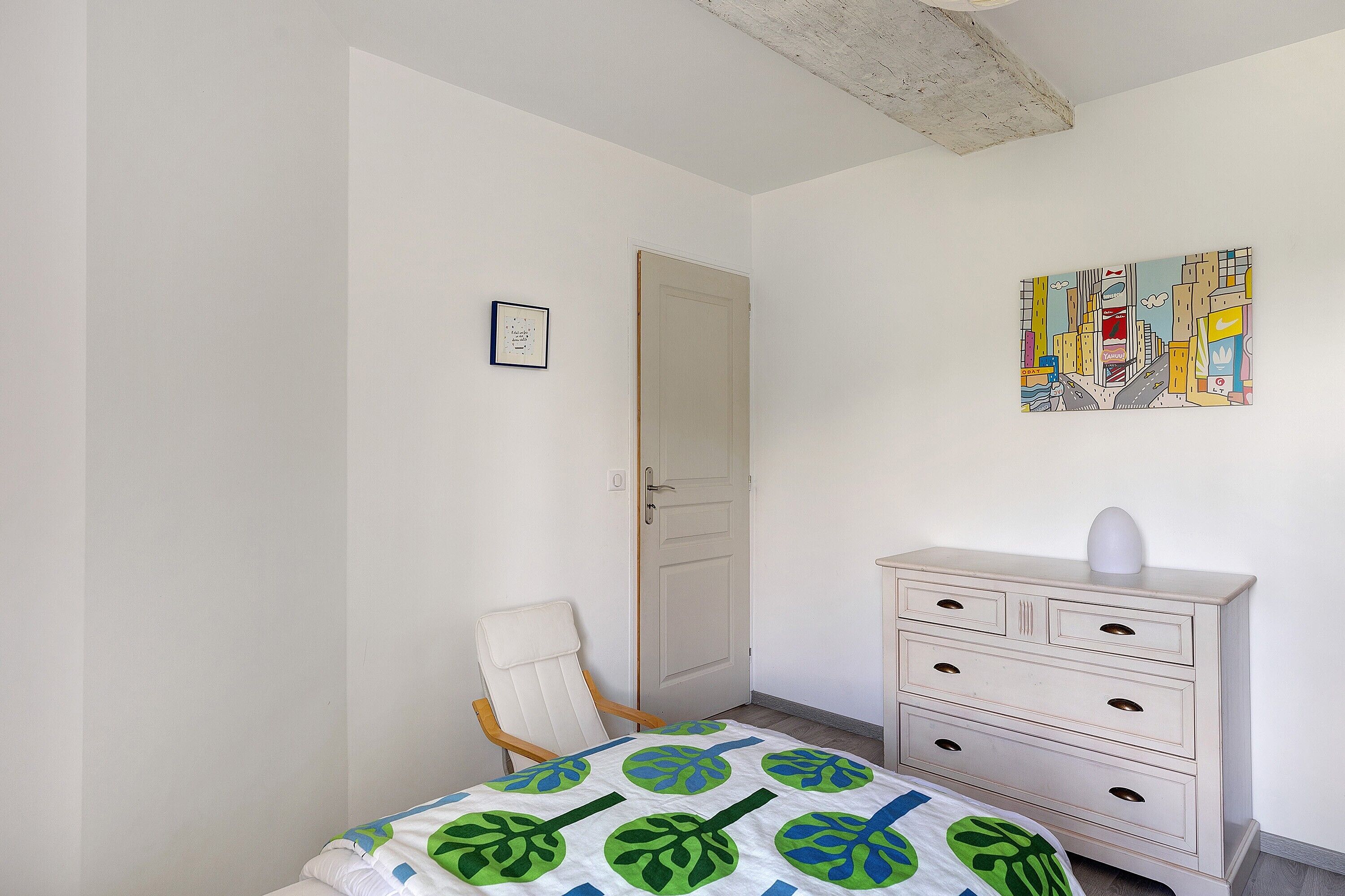 A bedroom with a bed, a chair, a dresser, a door, and a wall painting.