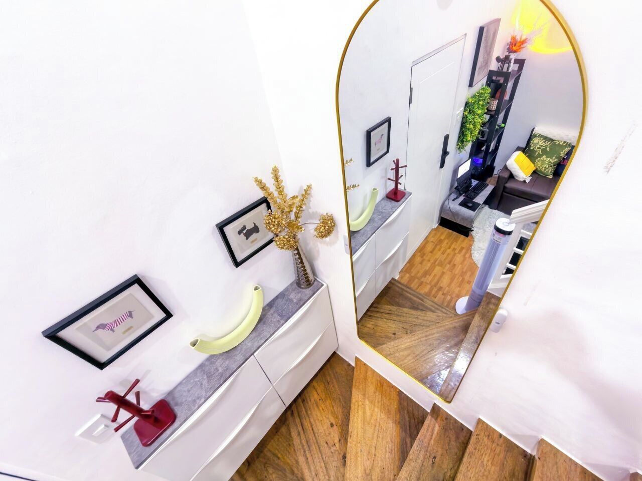 A modern bathroom with white walls, a wooden floor, and a large mirror. There are two sinks with white countertops and a small plant on the left sink.