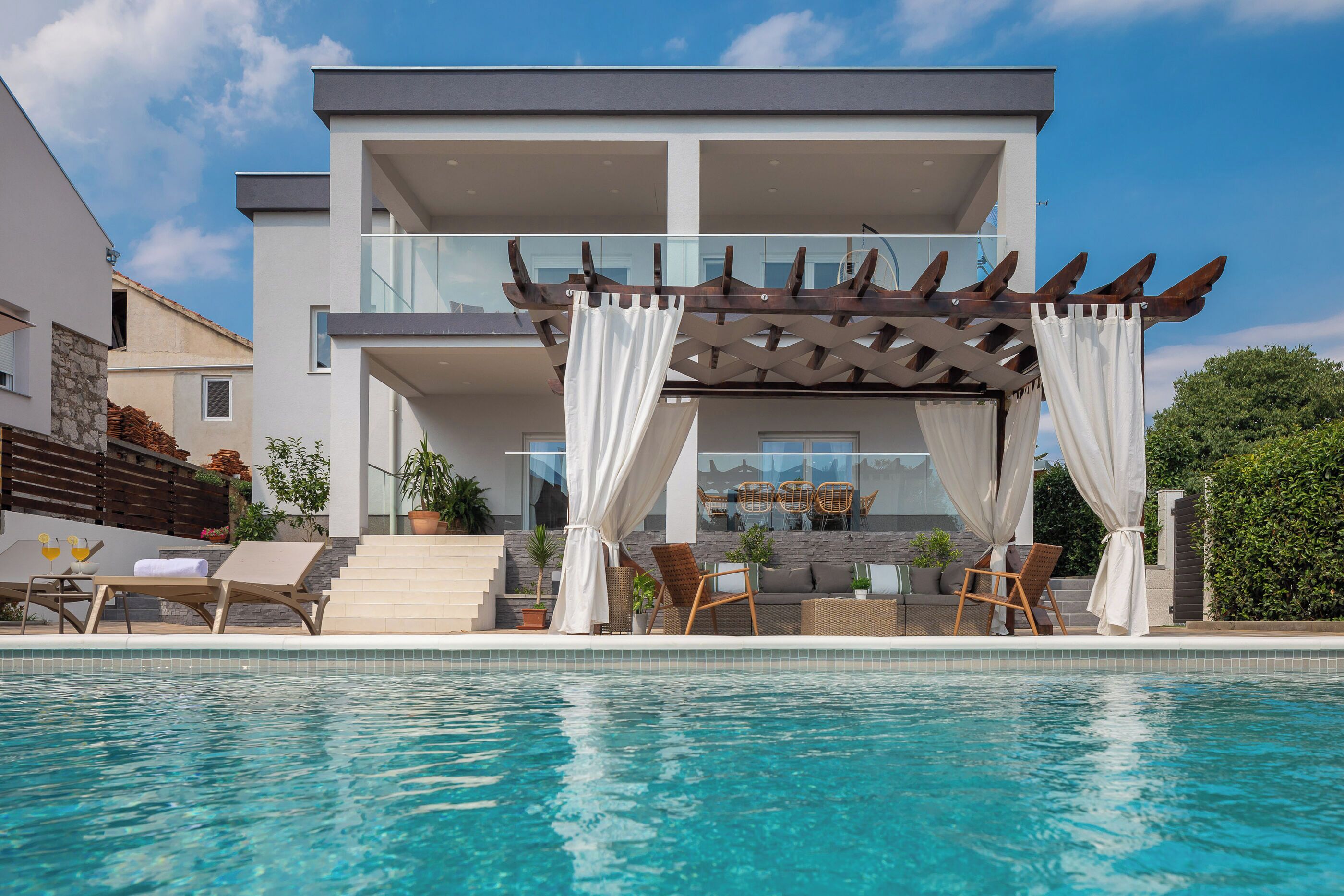 A modern two-story house with a pool, outdoor seating, and a pergola with curtains.