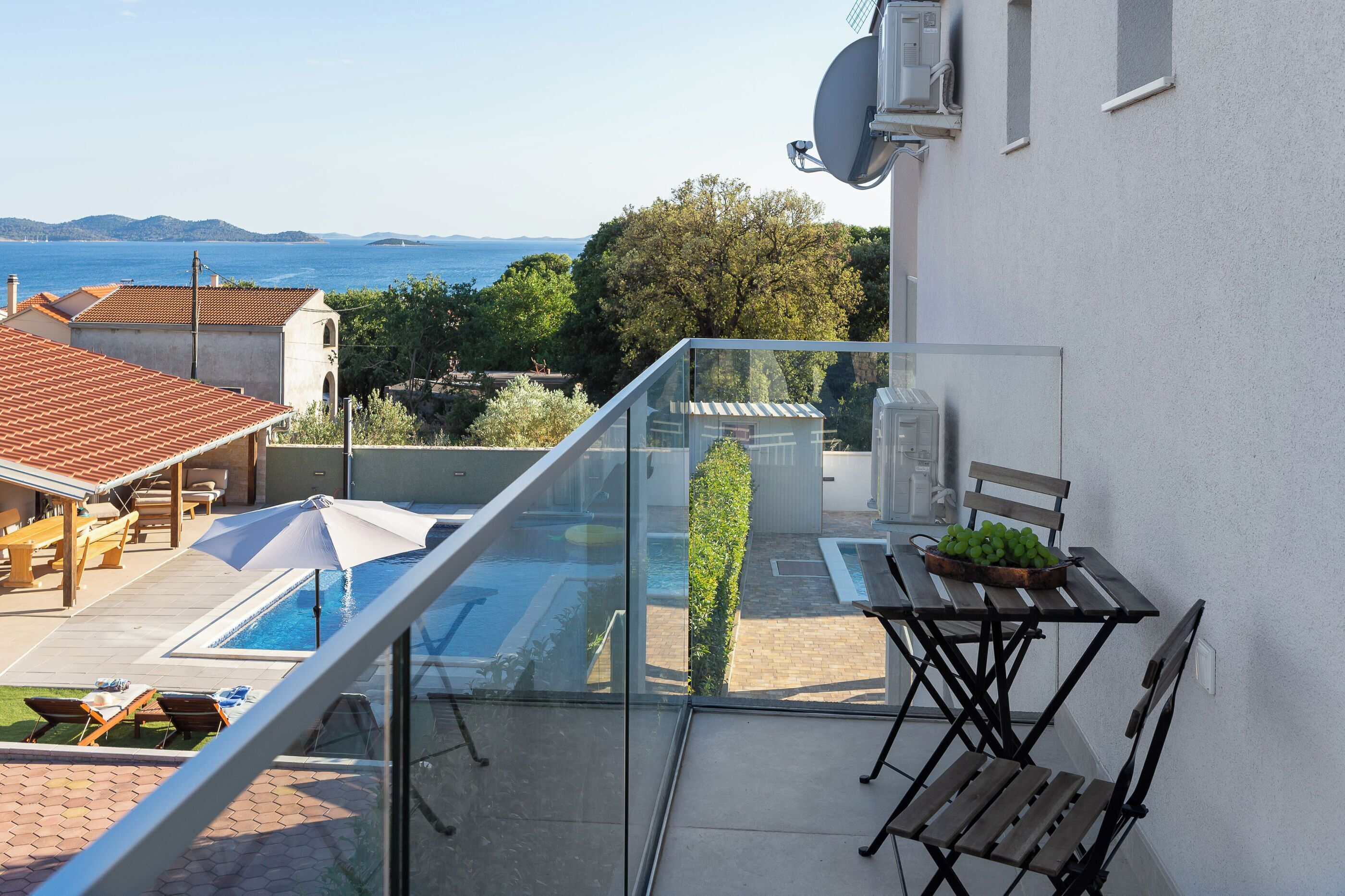 A balcony with a glass railing overlooking a swimming pool, a table with chairs, and a view of the sea and hills.