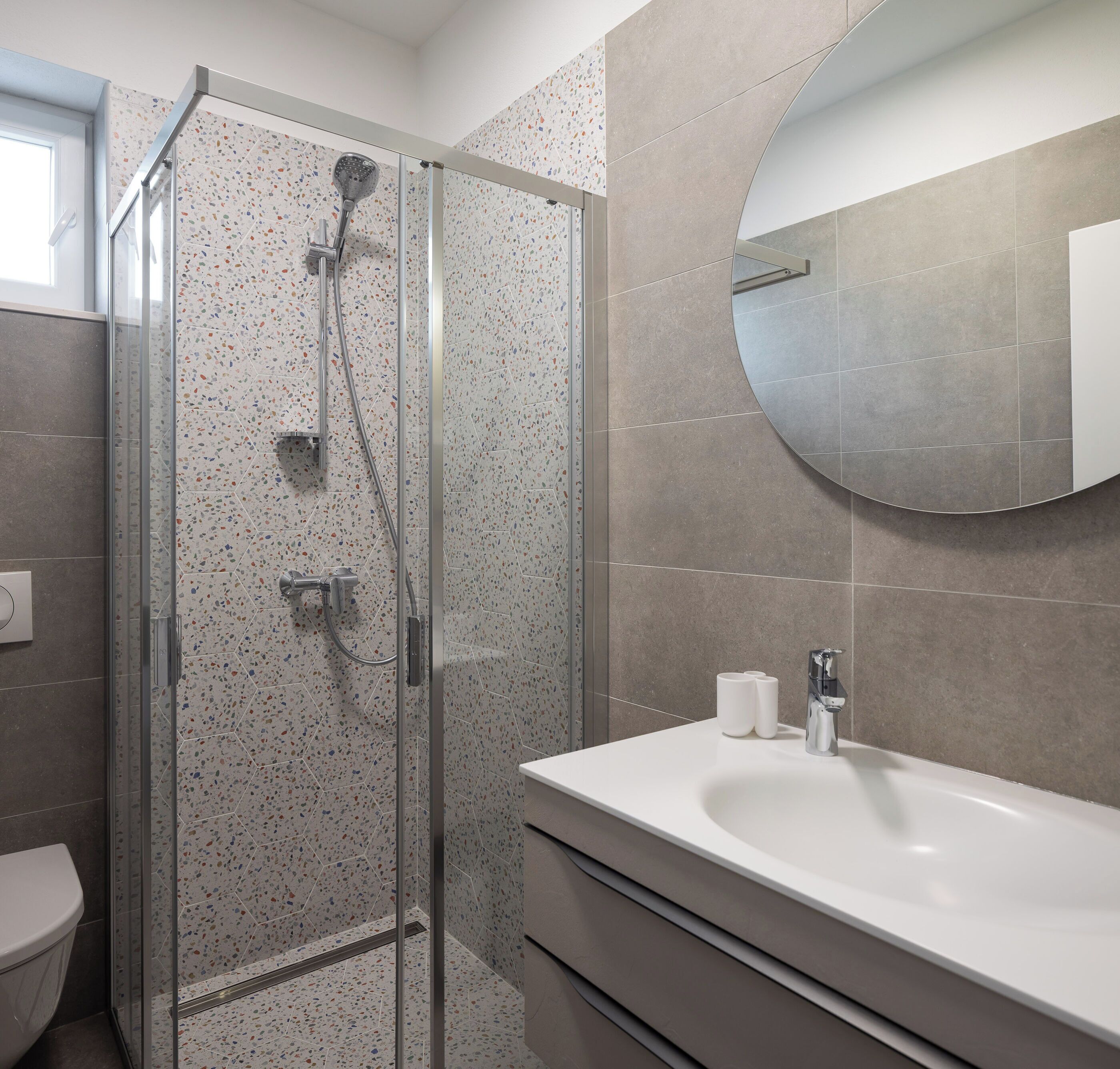 Egy modern fürőszoba üvegfalas zuhanyzóval, fehér mosdókagylóval és kerek tükörrel.
