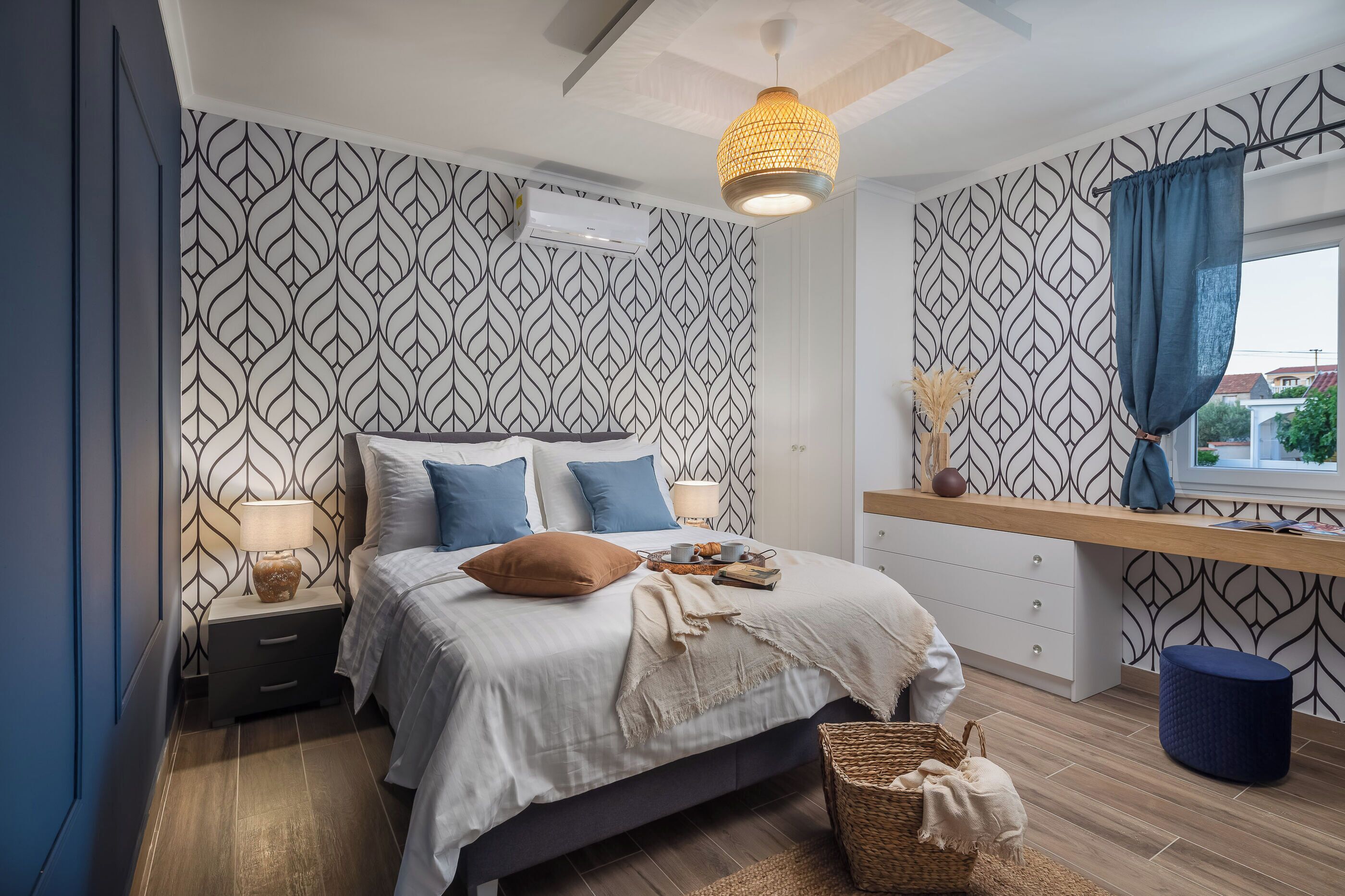 Quarto com papel de parede estampado, cama com almofadas azuis e castanhas, mesa de cabeceira com abajur e janela com cortina.