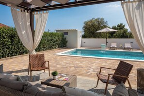 A poolside area with a pool, two wicker chairs, a small table with a book, and a covered seating area. - Villa Peregrine (Pakostane)