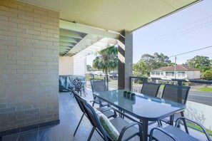 An outdoor patio with a glass table and chairs, a brick wall, and a view of the street and houses. - Spacious Flat With Parking & Dedicated EV Station (Batemans Bay)