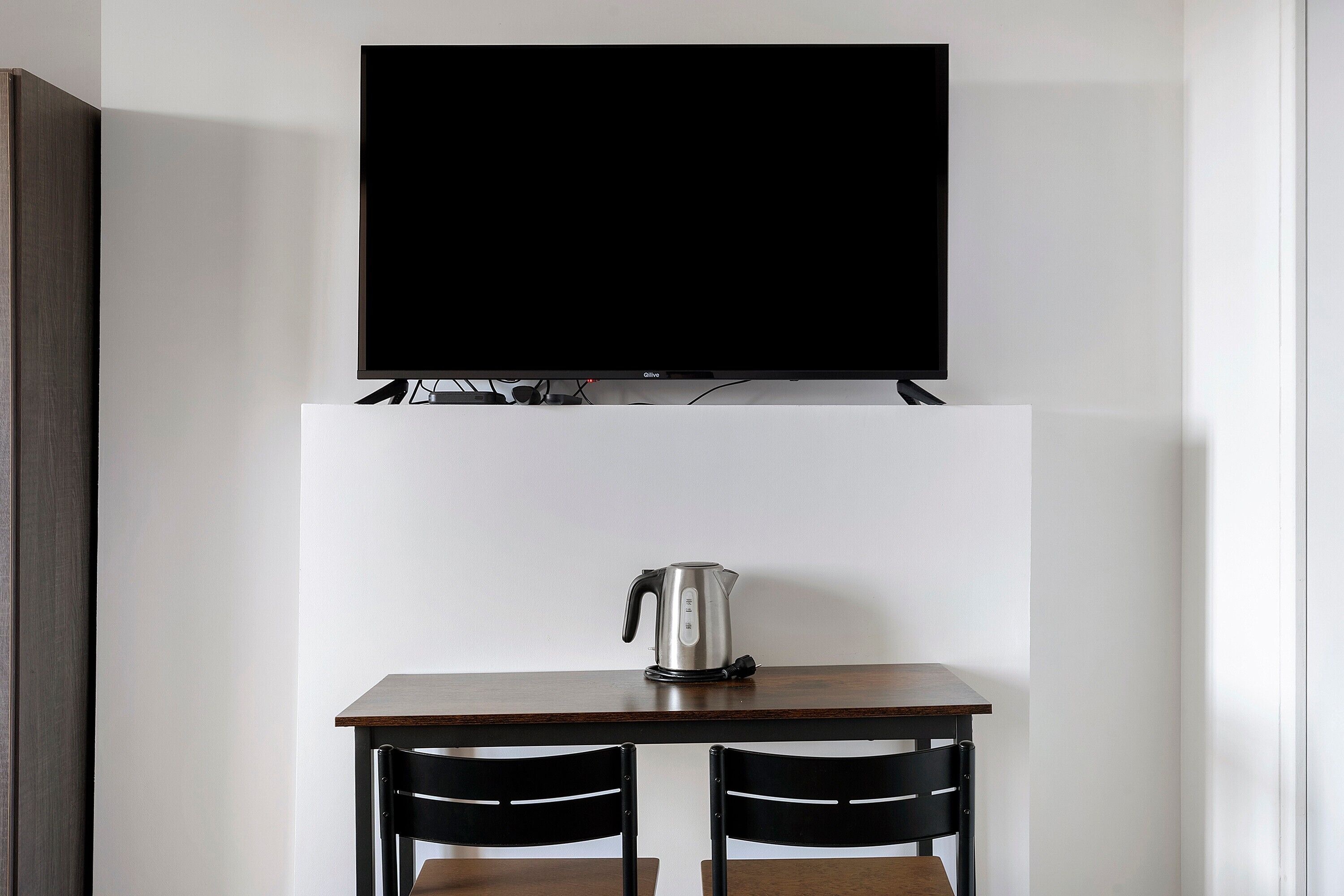 Quarto moderno com televisão de tela plana fixada acima de uma mesa de madeira com duas cadeiras, chaleira e tomada.