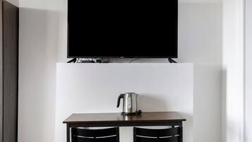 Quarto moderno com televisão de tela plana fixada acima de uma mesa de madeira com duas cadeiras, chaleira e tomada.