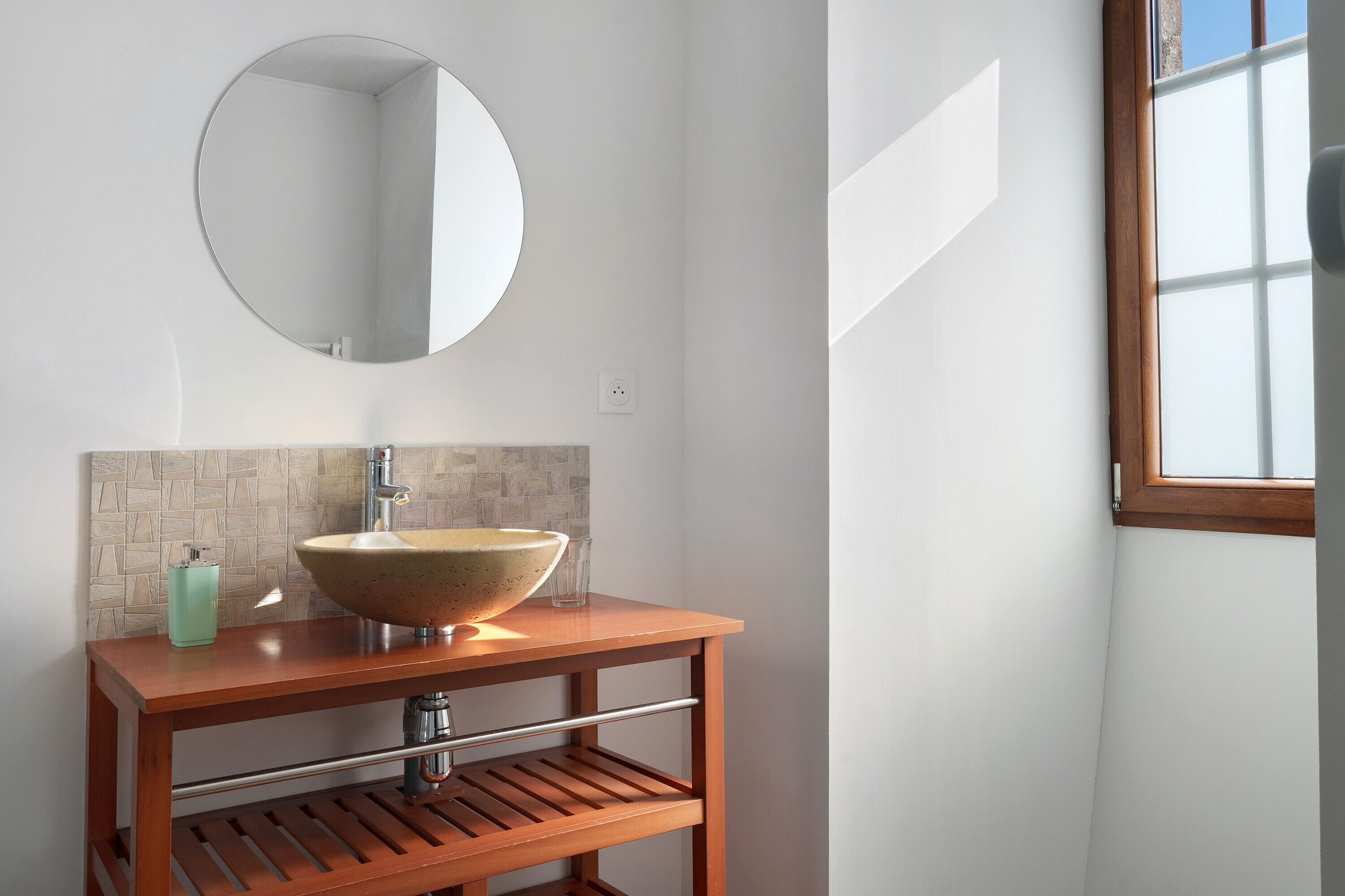 Une salle de bain avec un miroir rond, un meuble-lavabo en bois, un lavabo beige et une fenêtre avec un cadre en bois.