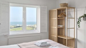 Ein Schlafzimmer mit großem Fenster mit Blick auf das Meer, einem Bett mit ordentlich gefalteten Handtüchern und einem Holzschrank mit Kleiderstange.
