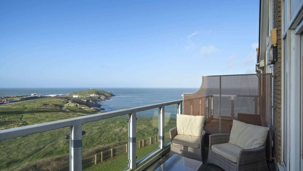 Ein Balkon mit Korbstuhl und Kissen mit Blick auf eine Küstenlandschaft, einschließlich Gebäude und einem Leuchtturm.