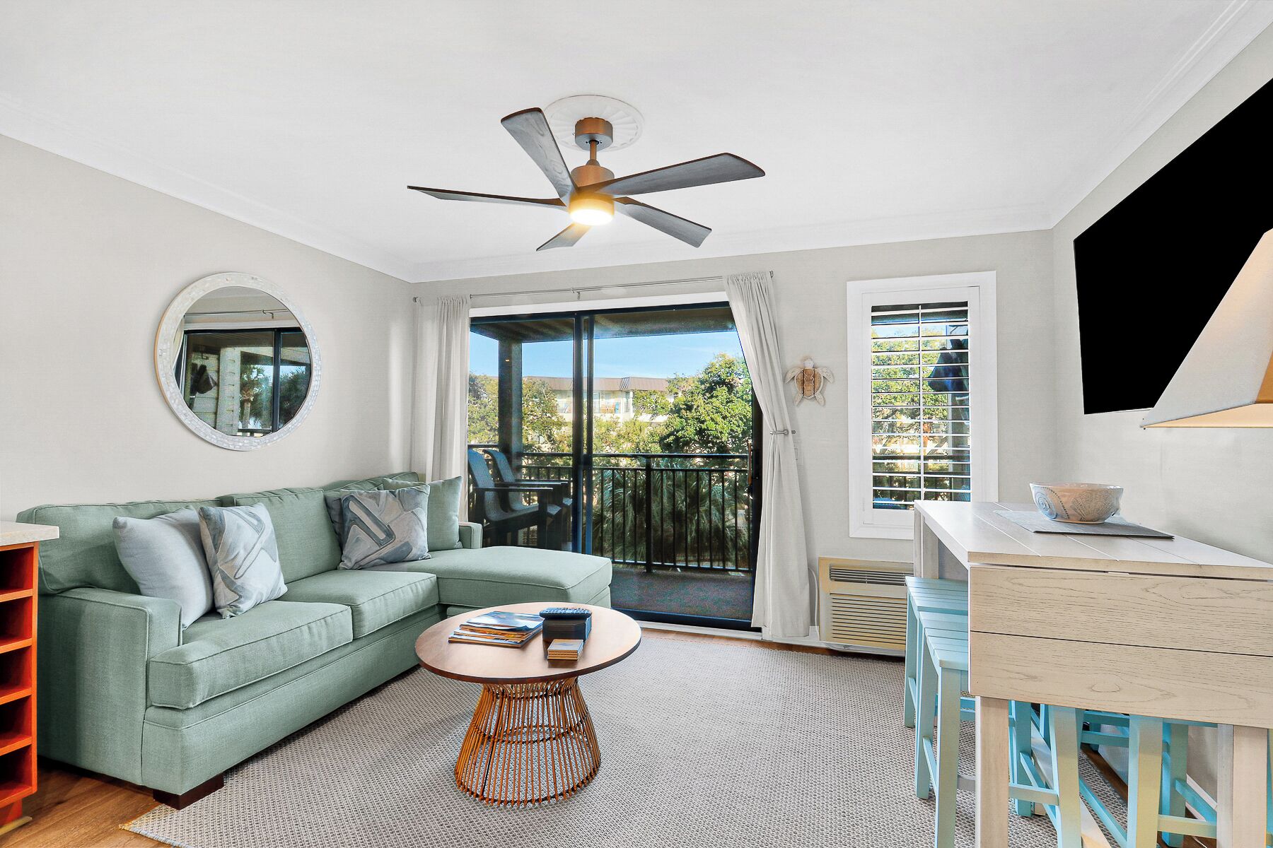 A living room with a green sofa, a round coffee table, a ceiling fan, and a view of the outdoors through a sliding glass door.
