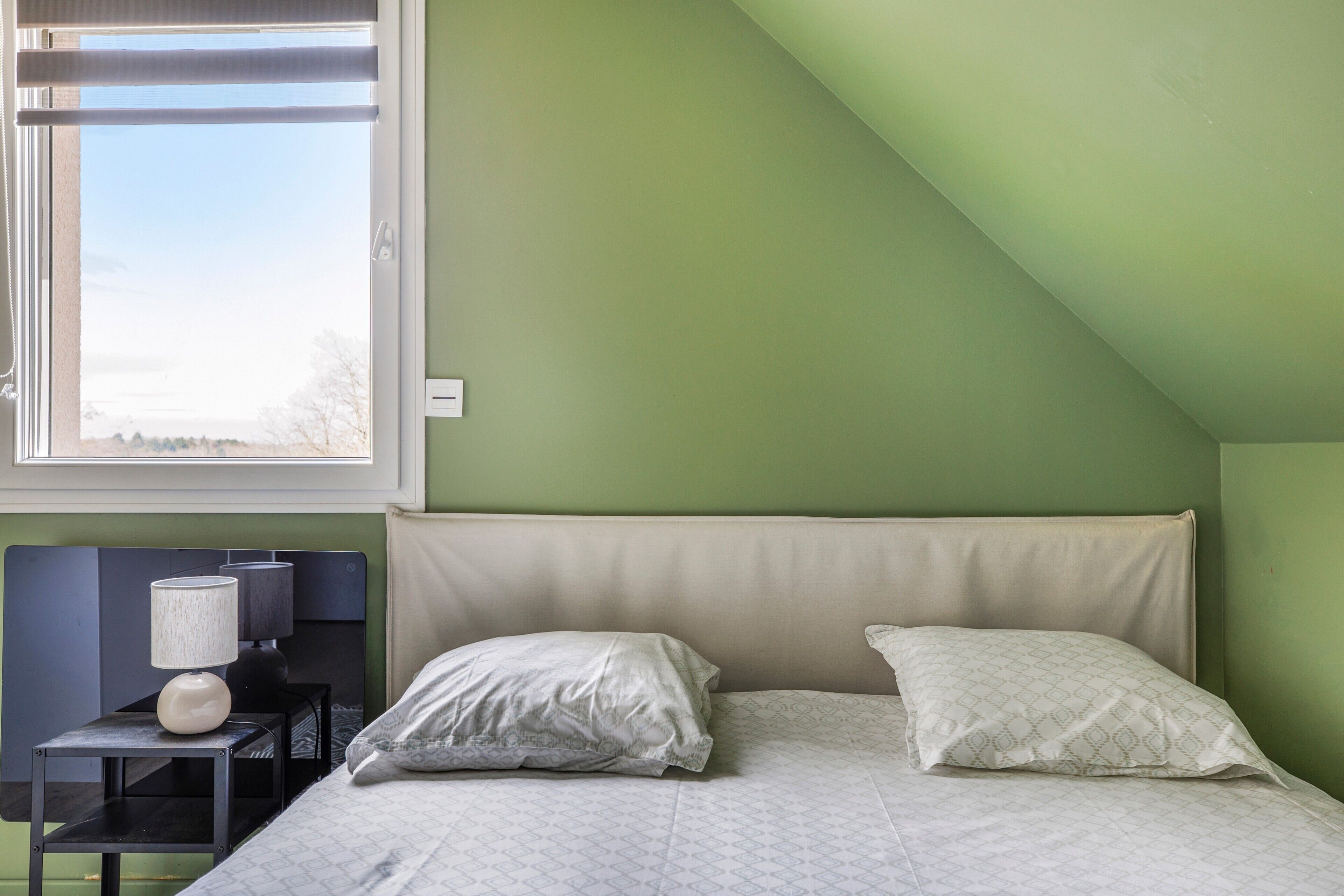 A bedroom with a bed, pillows, a nightstand, a lamp, and a window with a view.