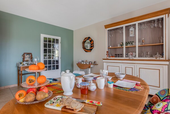 A dining area with a wooden table set for breakfast, a window with blinds, and a kitchen area with shelves and glassware. - Guest Room “La Gâtine” with Pool, Terrace & Wi-Fi in Rochecorbon (Rochecorbon)