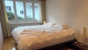 Un lit bien fait, avec du linge de lit blanc et une serviette pliée, une table de chevet avec une lampe, et une vue sur un bâtiment à travers une fenêtre avec des rideaux.